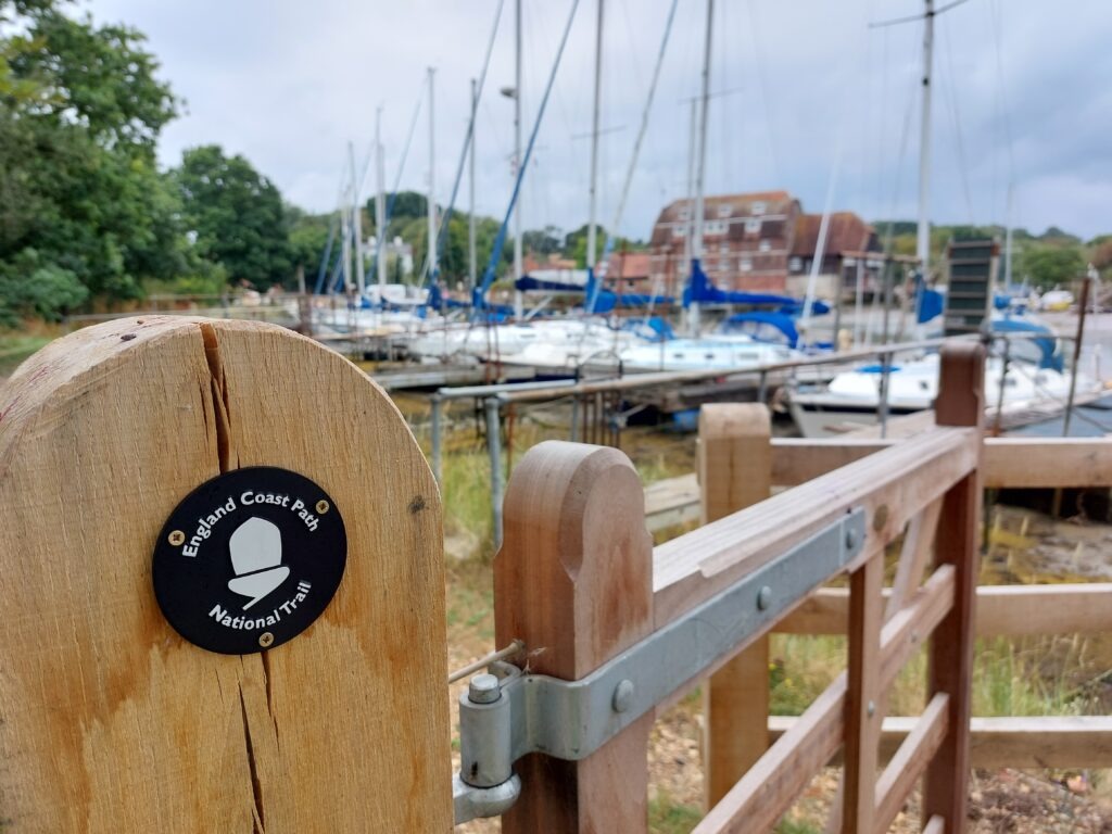 A gate with a badge on saying England coast path, with boats behind