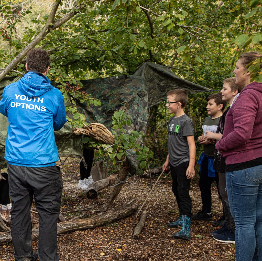 A group of children and a staff member from Youth Options building a den in a wooded area