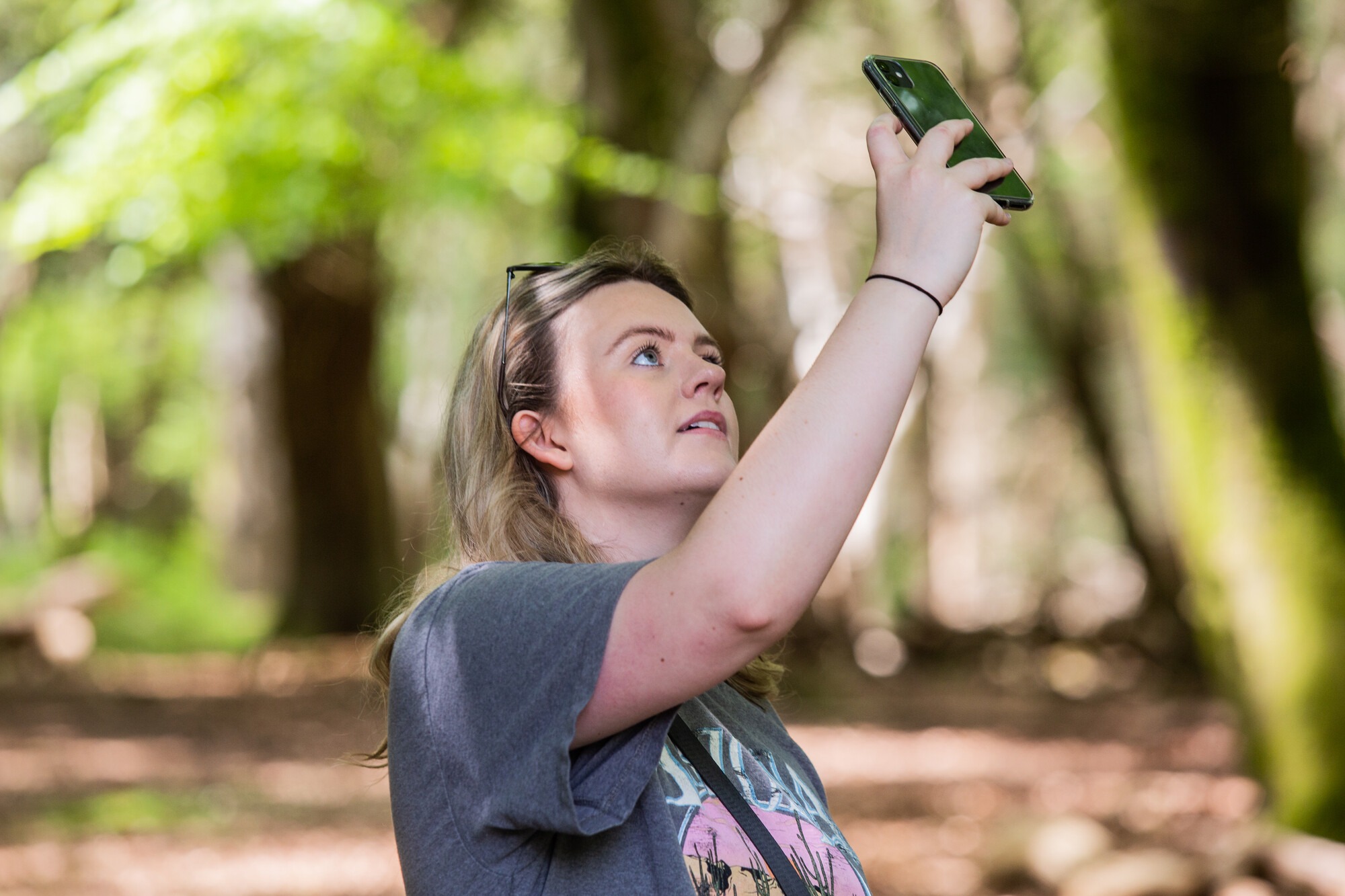 A woman holding a phone up to take a picture. She is stood in a wooded area.