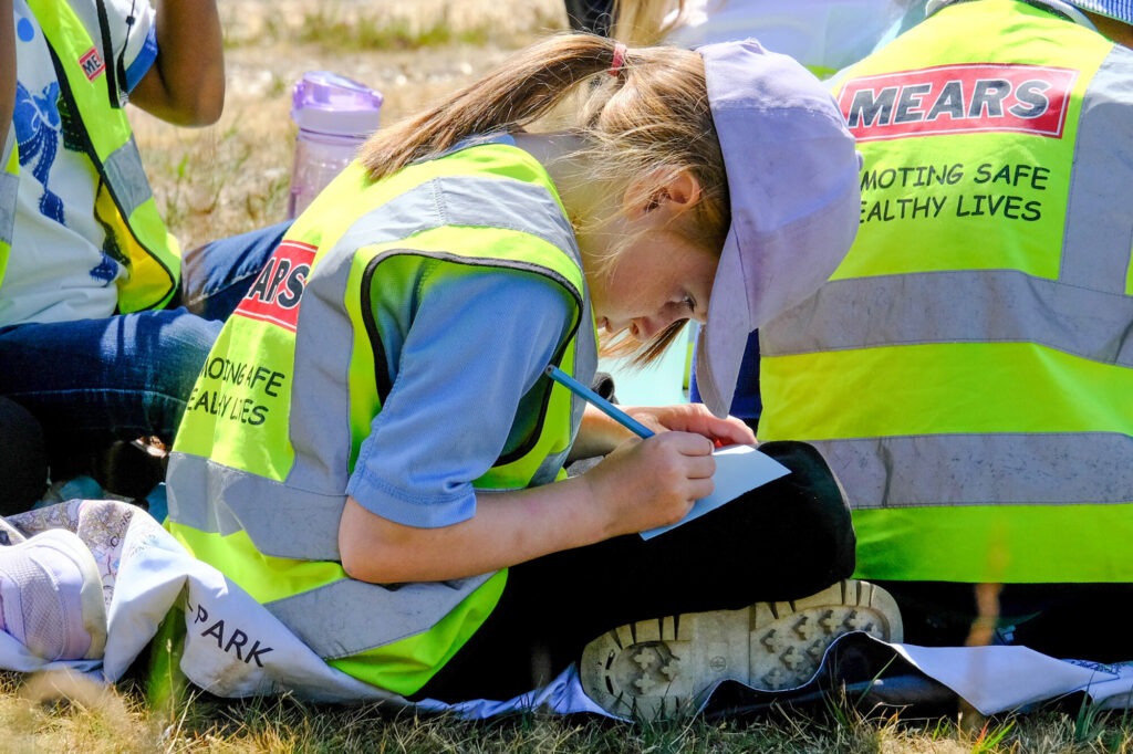A young school girl sat cross-legged on some grass writing on a bit of paper resting on her knee. She is wearing a hat and a high-vis jacket. It's a sunny day