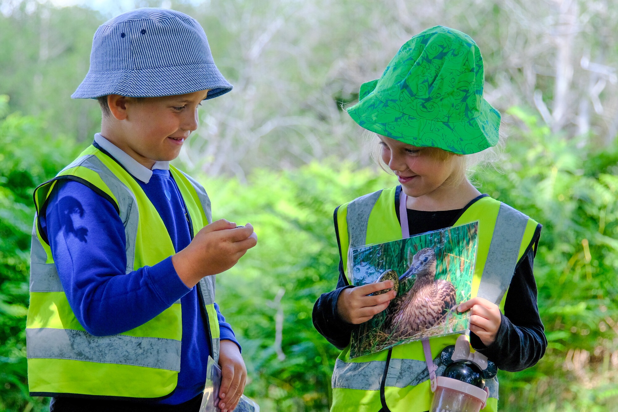 Two school children outside on a sunny day. They are both wearing hats and high-vis. They are on a school trip. One is holding a picture of a ground nesting bird the other is holding a model of an egg.