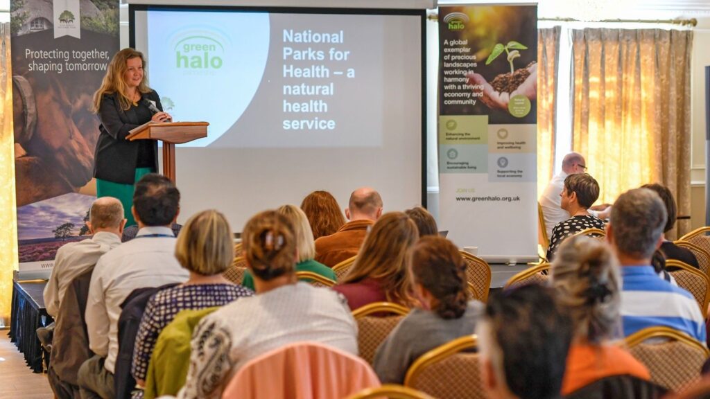 Alison Barnes speaks at a podium to an audience at Green Halo Conference 2024, beside a slide reading “National Parks for Health”