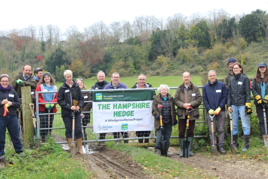 Hampshire Hedge Group volunteers with spades and gloves stand by a gate holding a “The Hampshire Hedge” CPRE banner in a field