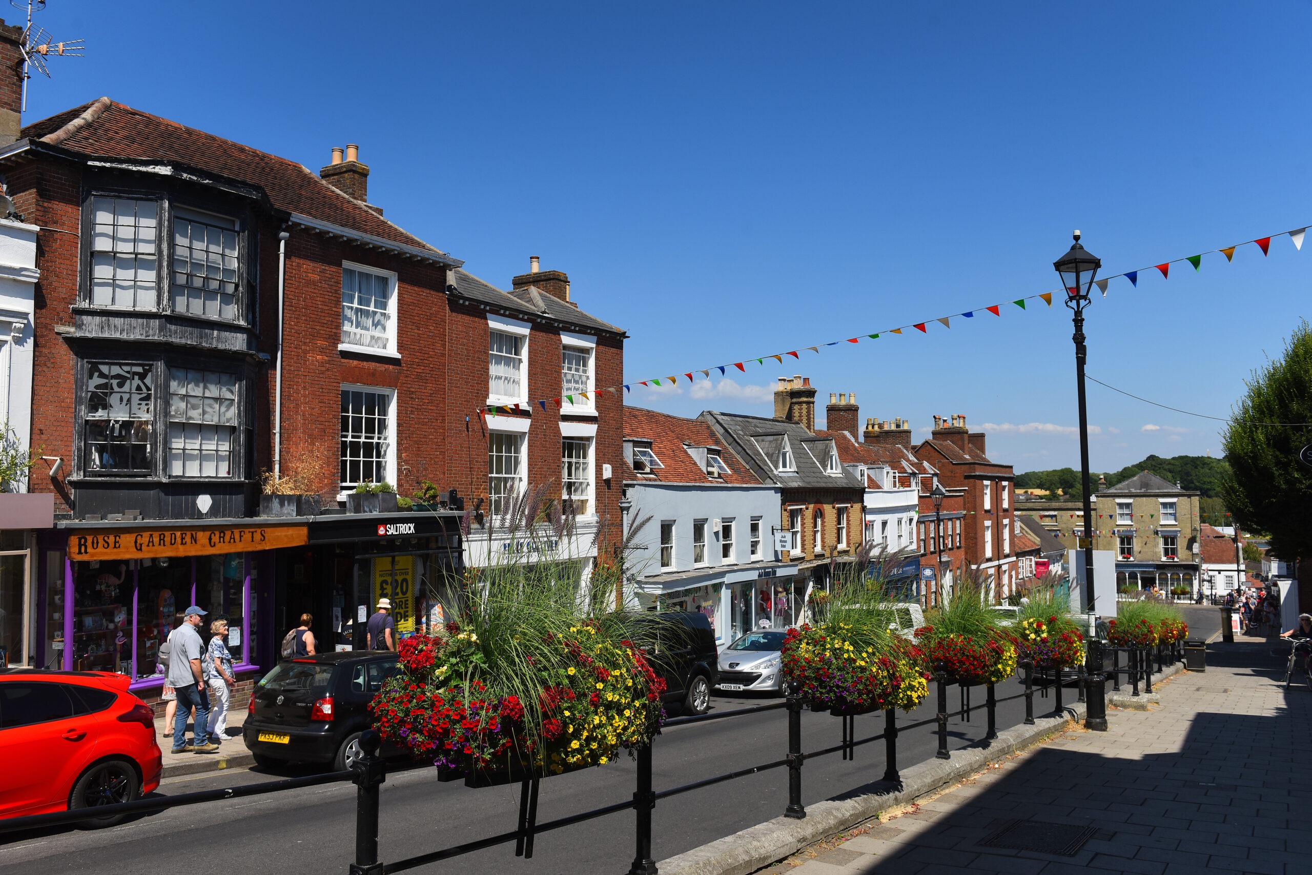 Lymington High Street with brick shops, hanging flower baskets, cars and pedestrians, and bunting across the road under blue sky