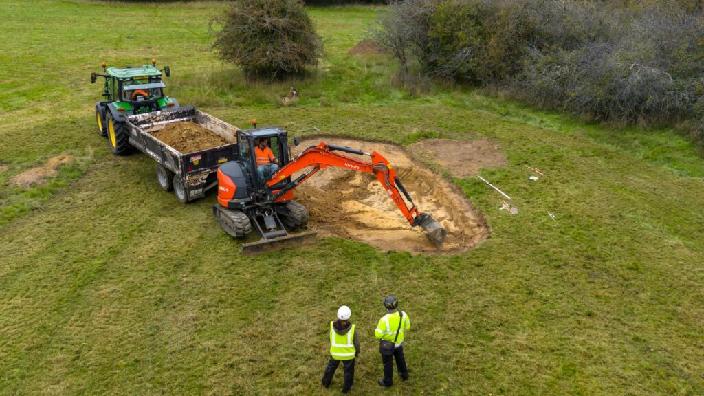 Two people watching a digger scraping out grass for a pond