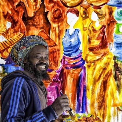 Smiling artist with paintbrush and palette in front of a colorful painting of tall figures in warm orange and purple tones