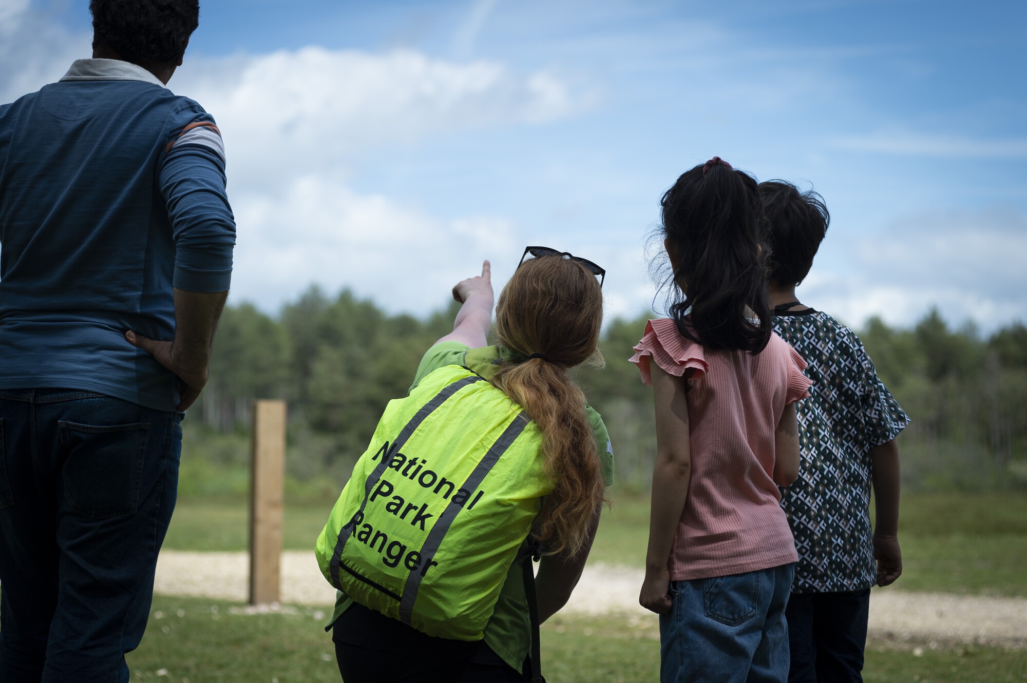 A woman wearing a fluorescent New Forest ranger backpack crouches beside two children and points to something in the distance. An adult man is stood next to her. They are in a grassy area with trees in the distance