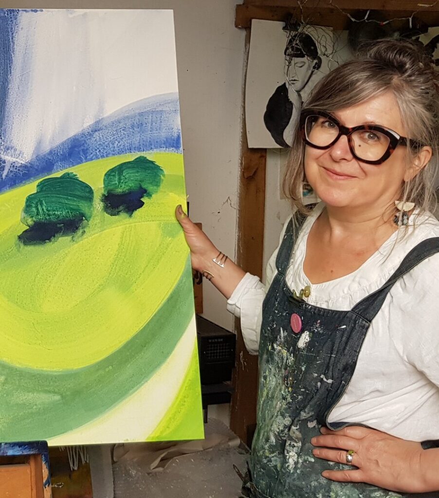 Artist in paint-splattered apron holds a canvas showing a green landscape with two trees in a studio