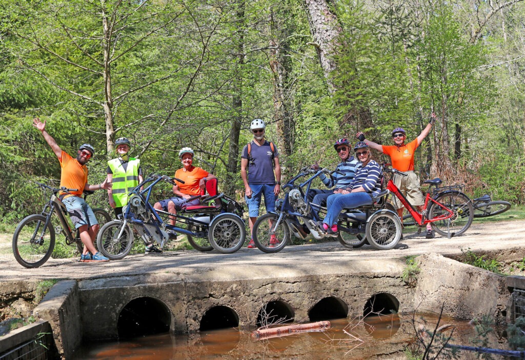 Seven riders stand and sit on a range of bikes and specialised cycles on a ride in the New Forest in a wooded area.