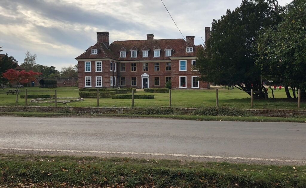 Large red-brick manor house with dormer windows and chimneys behind a fenced lawn, viewed from across a road under cloudy sky