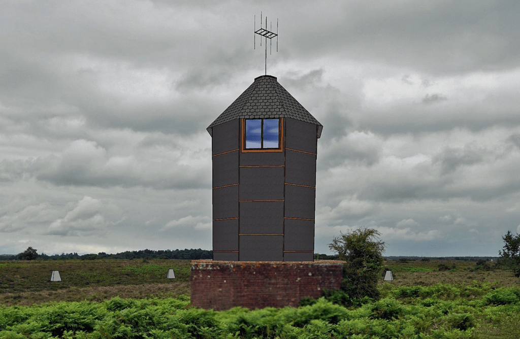 Small hexagonal lookout tower with a conical roof and antenna, standing on a brick base in heathland under cloudy sky