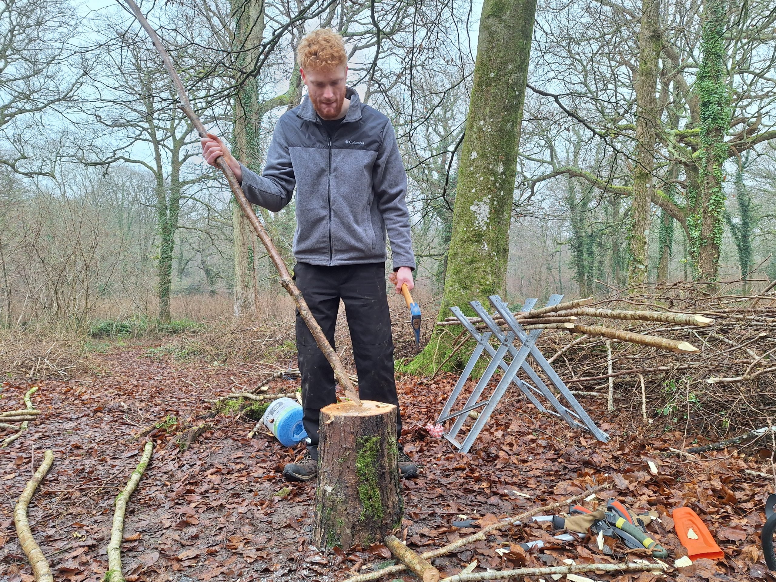 CET intern Charlie holding an axe in one hand and slim coppiced tree in the other, he is about to chop one end into a fine point