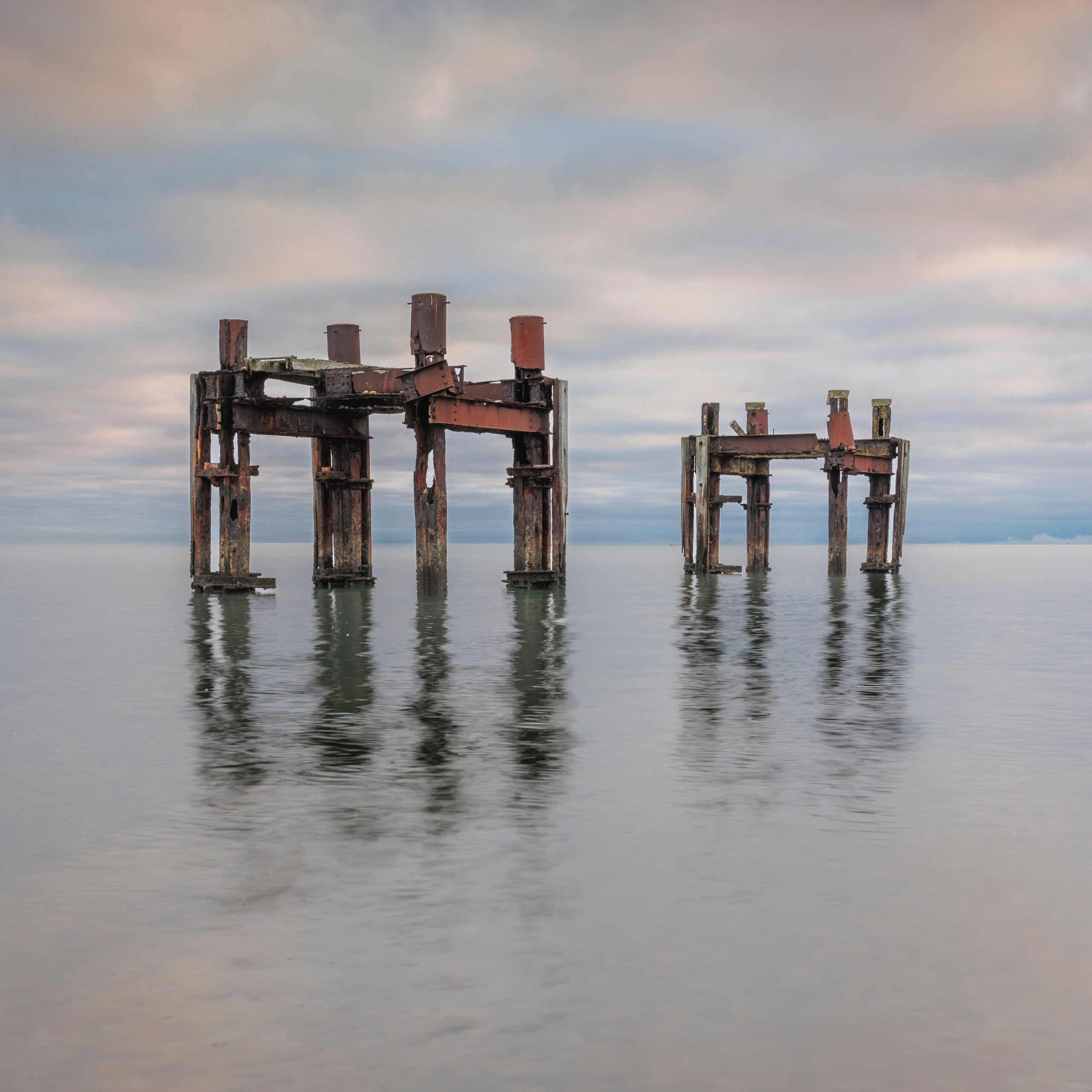 Two rusted WWII loading pier dolphins standing in calm sea at Lepe Beach, Hampshire, reflected under a cloudy sky
