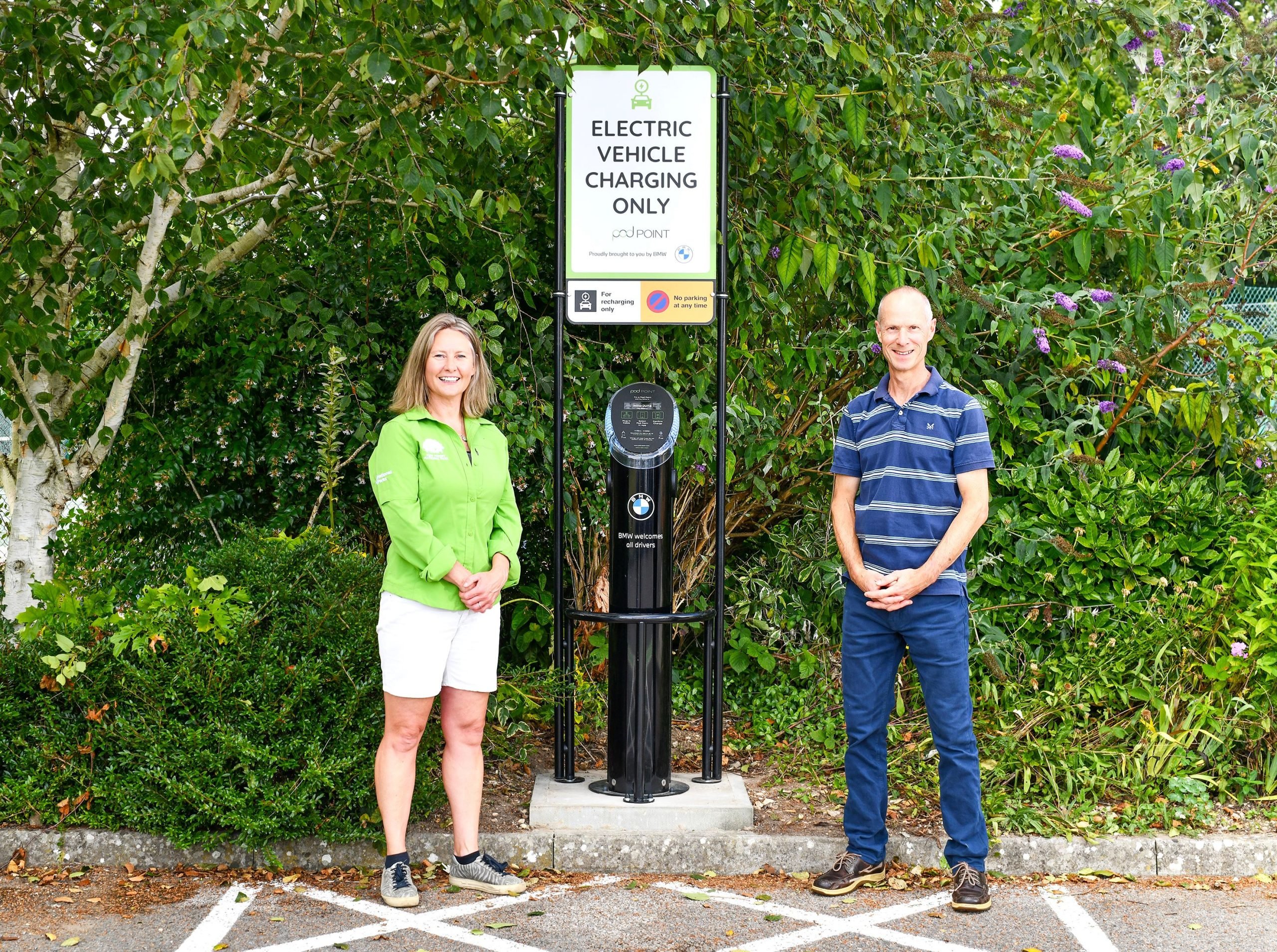 A man and woman stand either side of an EV electric charging point with trees behind