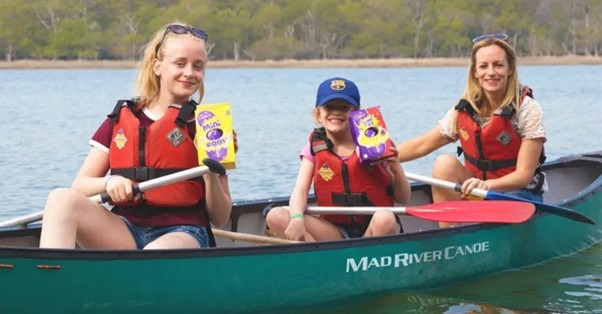 Three people in a canoe wearing life vests, holding Cadbury Creme Egg boxes on a lake