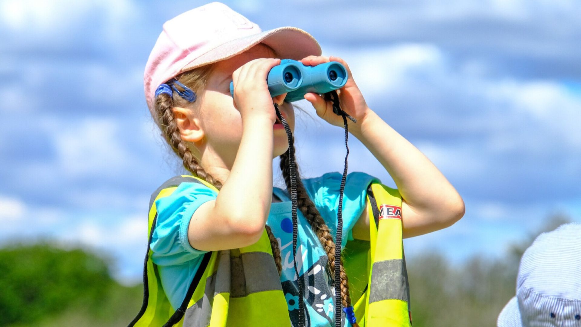 Child in a pink cap and safety vest looks through blue binoculars outdoors against a cloudy sky
