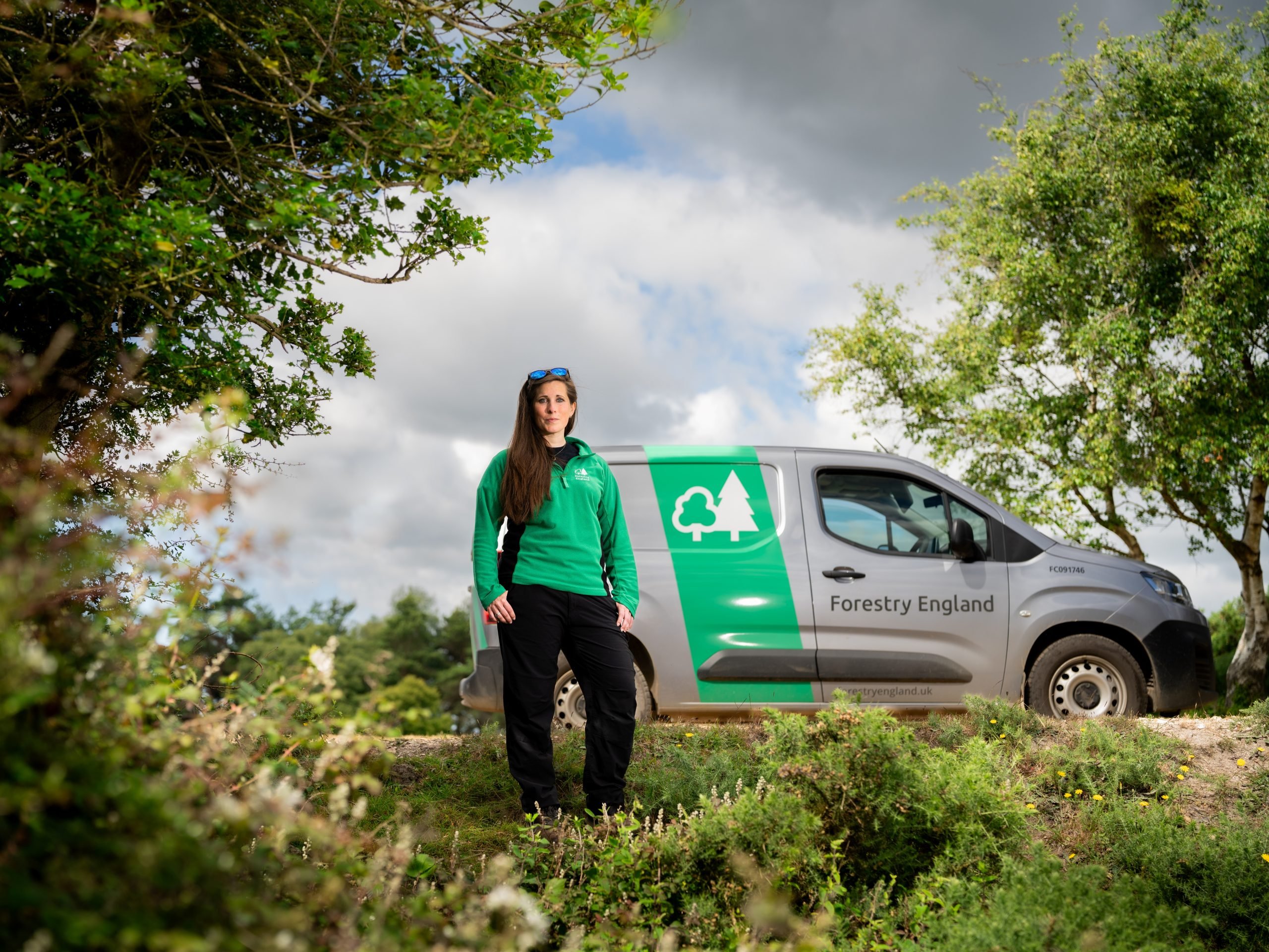 Forestry England ranger Charlotte Belcher standing by a Forestry England van in woodland under a cloudy sky