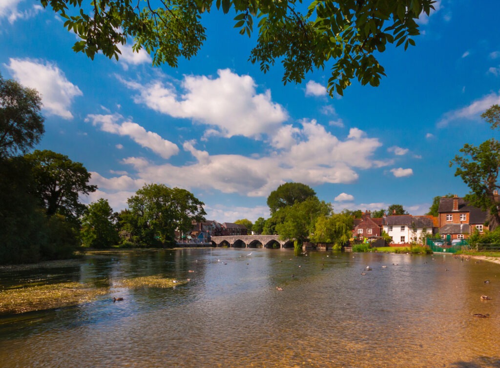 River Avon at Fordingbridge, Hampshire, with stone arch bridge, riverside trees and houses under a blue sky with clouds