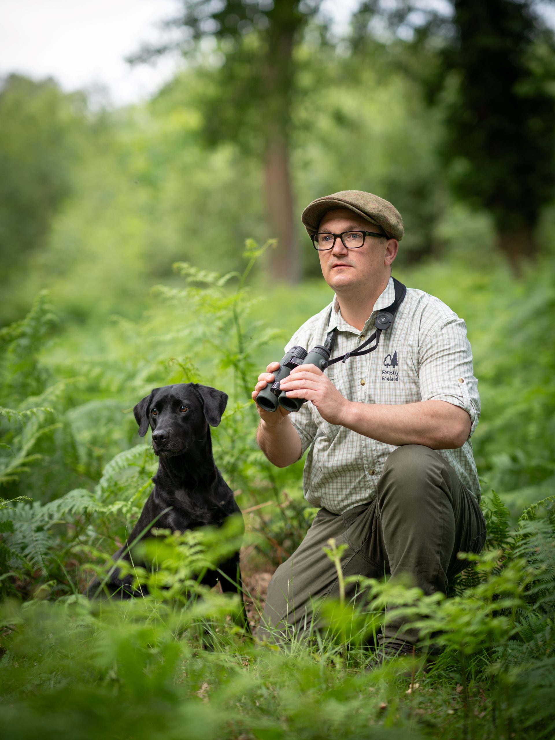 Forest Keeper Lee Knight crouched in woodland with his dog beside him, he is holding binoculars and looking off into the distance