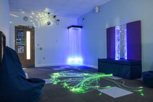 Sensory room at Brockenhurst College with bubble tube, fiber-optic lights on floor, and hanging light strands casting colored dots on walls