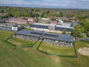 Aerial view of Brockenhurst College campus with modern buildings, solar panels on roof, sports field in foreground, and trees beyond