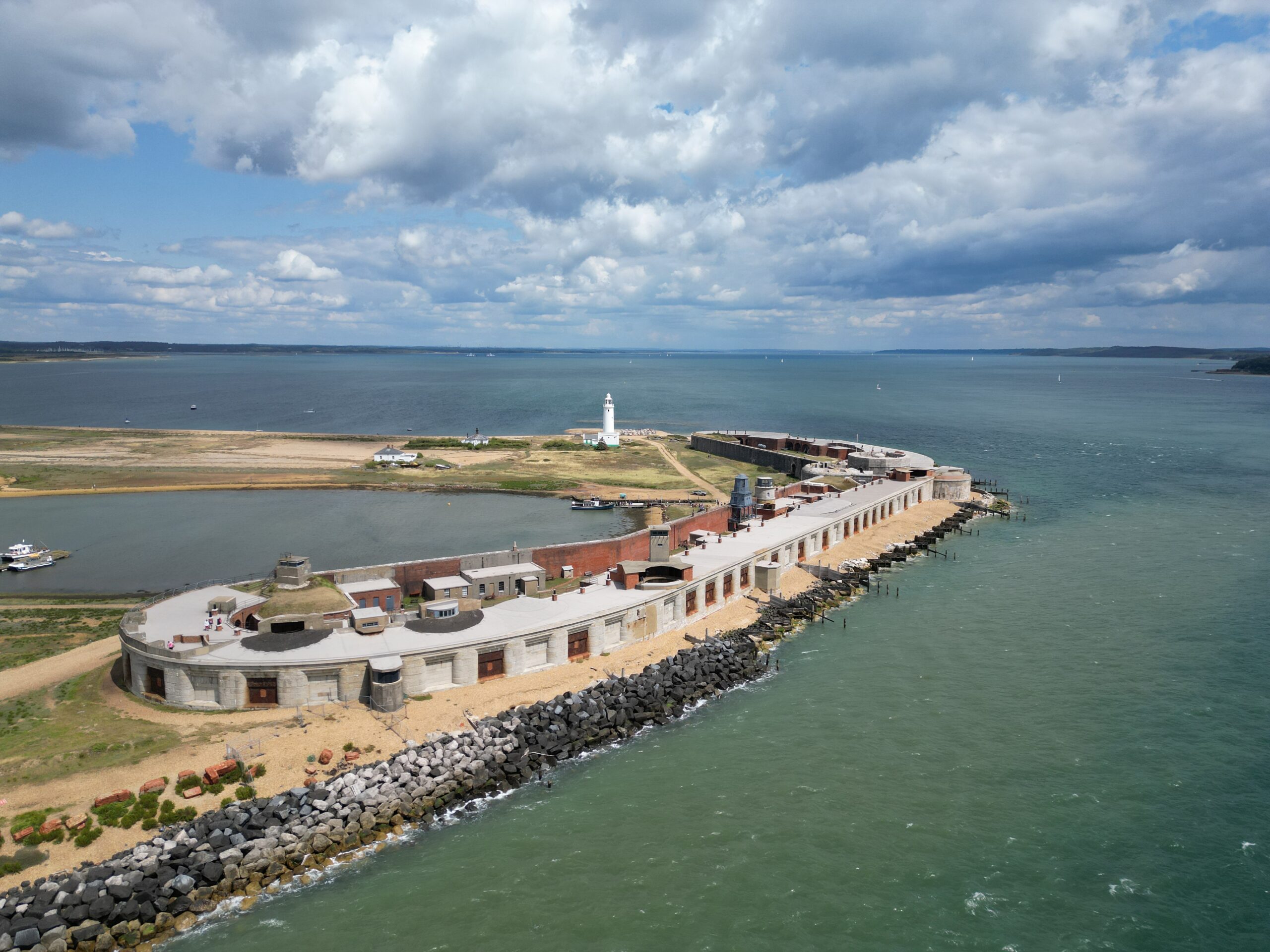 Drone view of Hurst Castle on a narrow shingle spit, with a lighthouse and sea on both sides under cloudy sky
