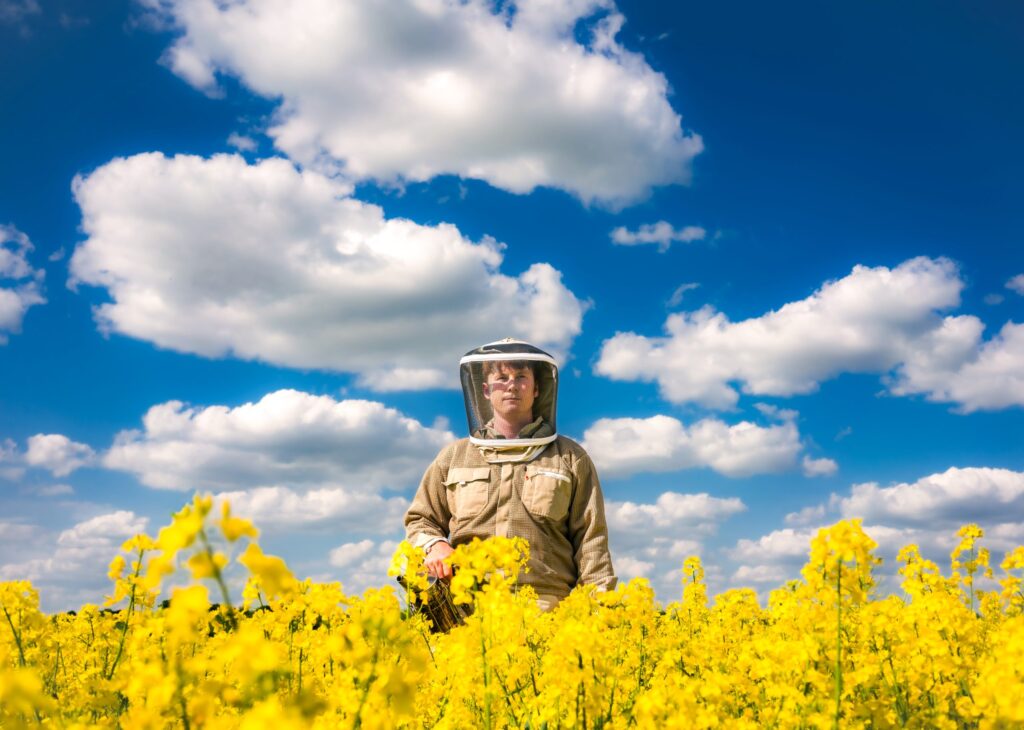 Beekeeper Jack Stride in a bee suit stood in a field of yellow flowers on a bright sunny day