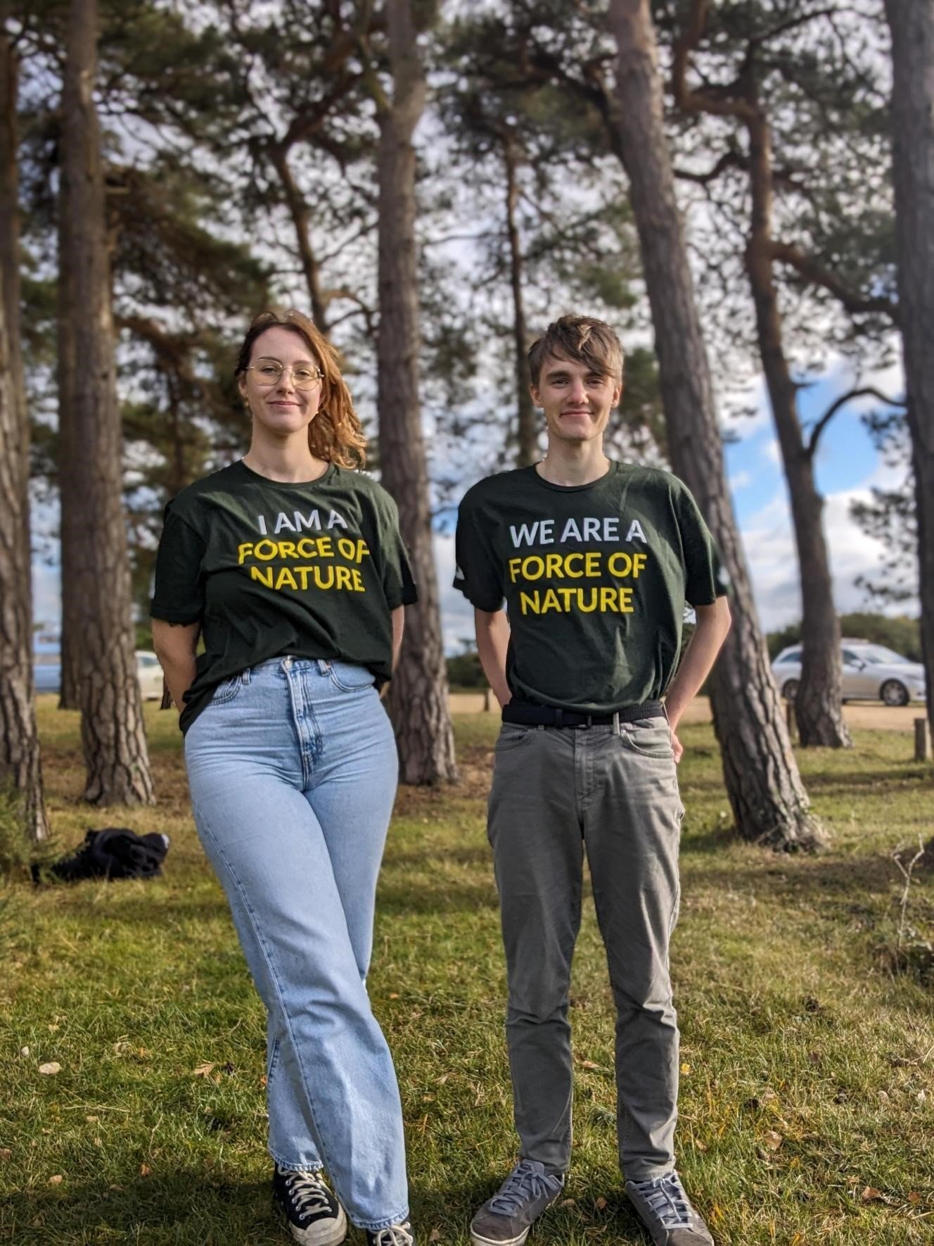 National Park interns Hayley on the left, Josh on the right. Both stood in a forest clearing with trees and blue sky behind. They are wearing green t shirts which read we are a force of nature