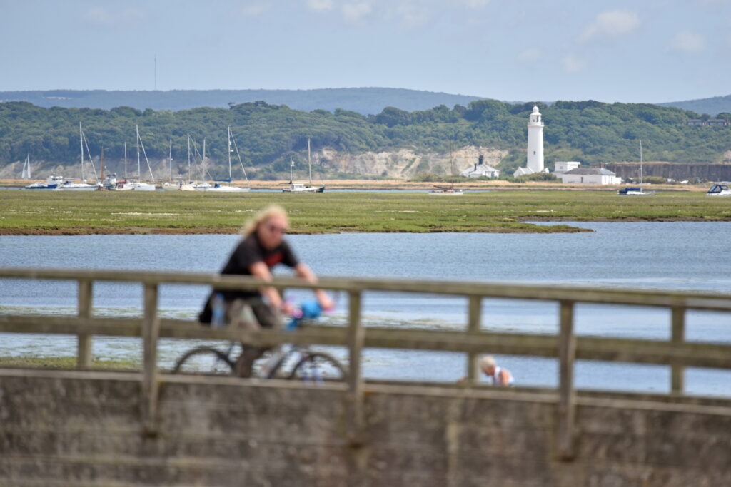 View across Milford-on-Sea harbour with sailboats and a white lighthouse, blurred cyclist riding along the seafront in foreground