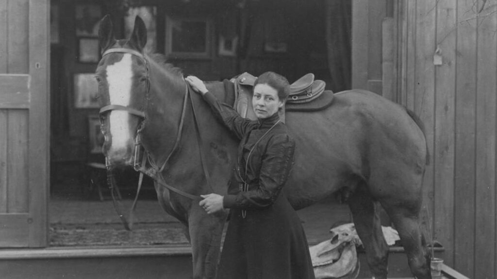Black-and-white photo of a woman standing beside a saddled horse, holding the reins outside a stable doorway