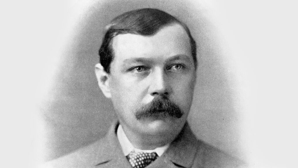 Black-and-white portrait of a mustached man in a suit and tie, facing slightly right against a plain background