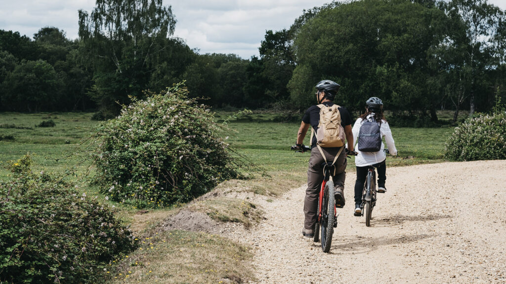 Two cyclists wearing helmets and backpacks ride on a gravel path through a green countryside with trees
