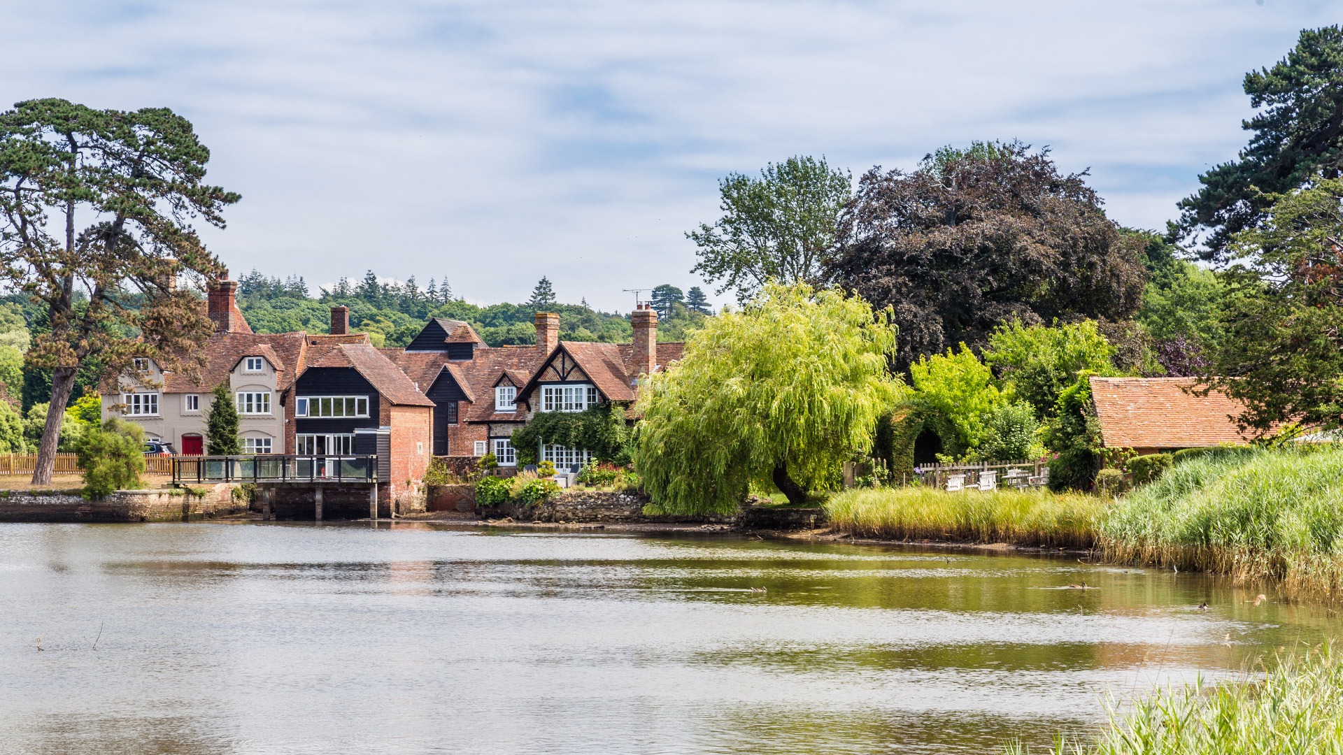 Riverside village houses with red-tiled roofs and chimneys, surrounded by trees and reeds along a calm river under cloudy sky