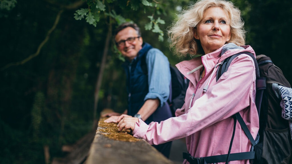Older woman in pink jacket with backpack leaning on wooden railing in forest, with smiling man in background
