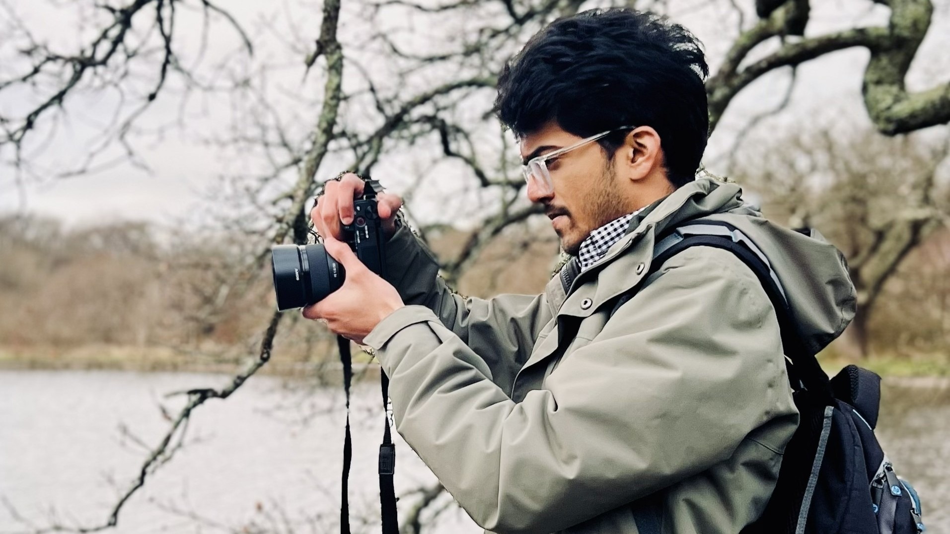 Omkar Dherange photographing by a lake under bare tree branches, wearing glasses, a green jacket and a backpack