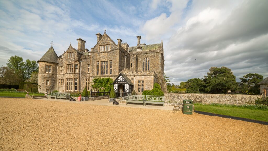 Palace House in Beaulieu, UK, a large stone manor with gables and turret, seen from the gravel courtyard under a cloudy sky