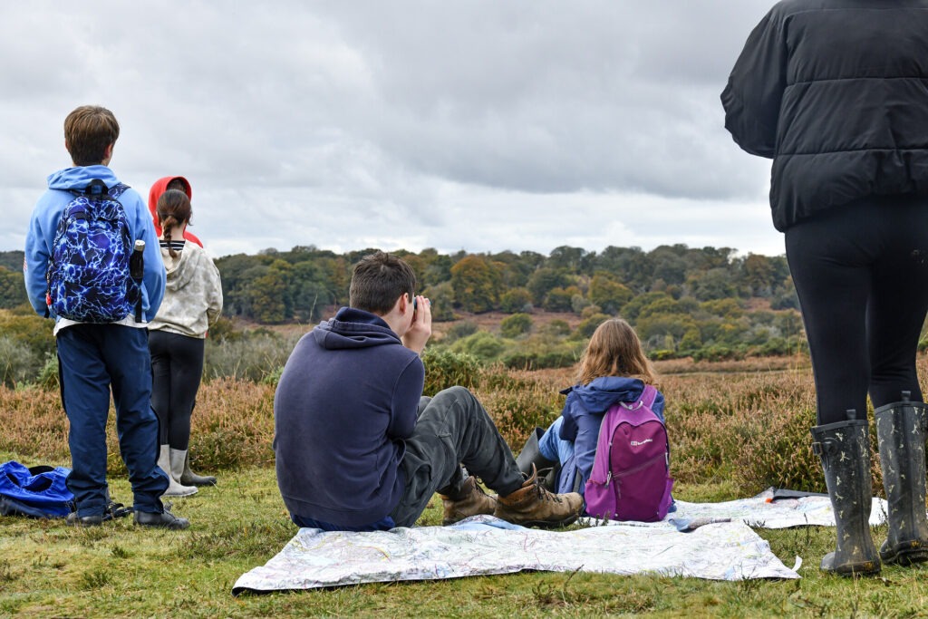 Some young people sat on a picnic blanket and others standing looking out at a viewpoint in the New Forest