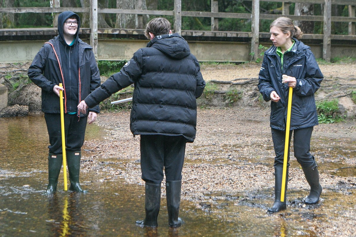NPA education officer stood by a riverside with two secondary school pupils in the shallow river. All are holding large rulers.