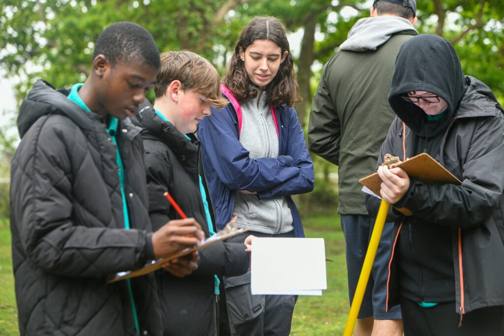 A group of teenage boys and a supervisor in a woodland area holding metre rulers and clipboards
