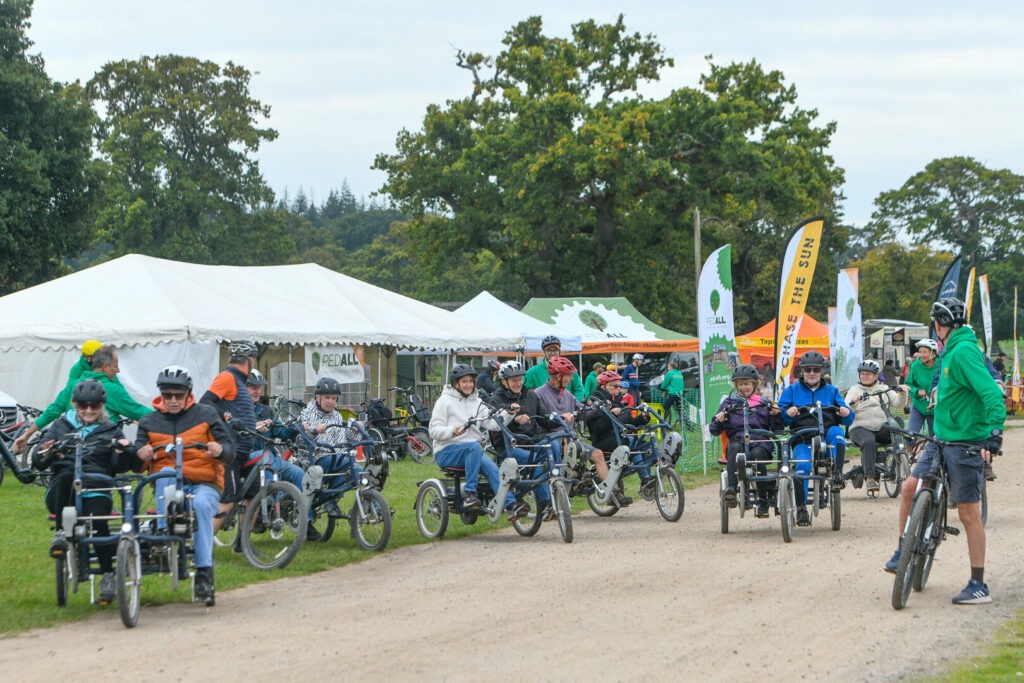 Riders on adapted cycles at Pass the PedALL event 2025 in New Park, Brockenhurst, with tents and flags behind