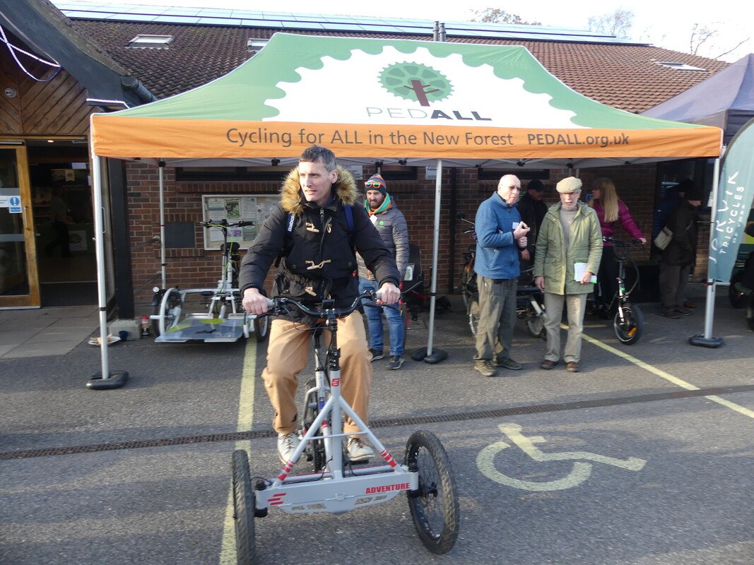A man on a recumbent tricycle in a car park. There is a PEDALL New Forest Inclusive Cycling marquee behind him