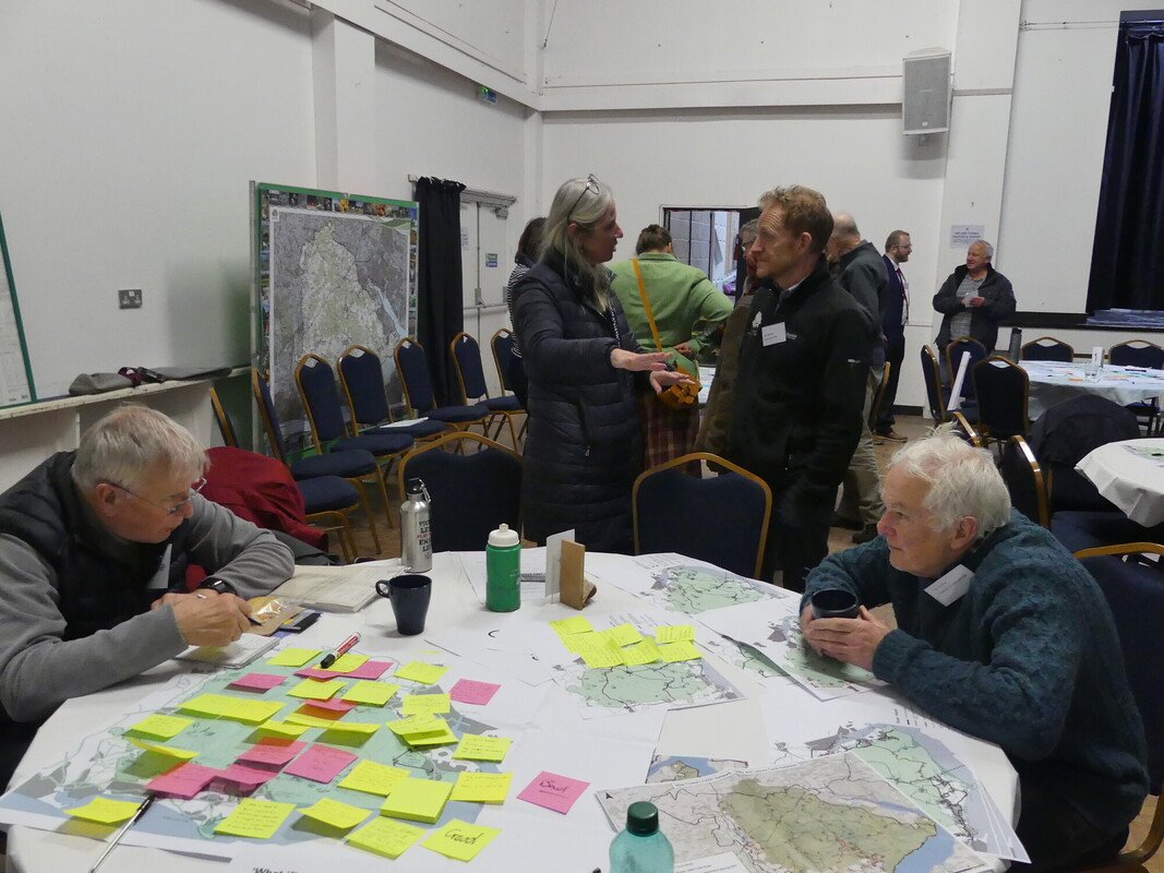 Two people sat at a table with maps of the New Forest and post it notes. Others are stood talking. 