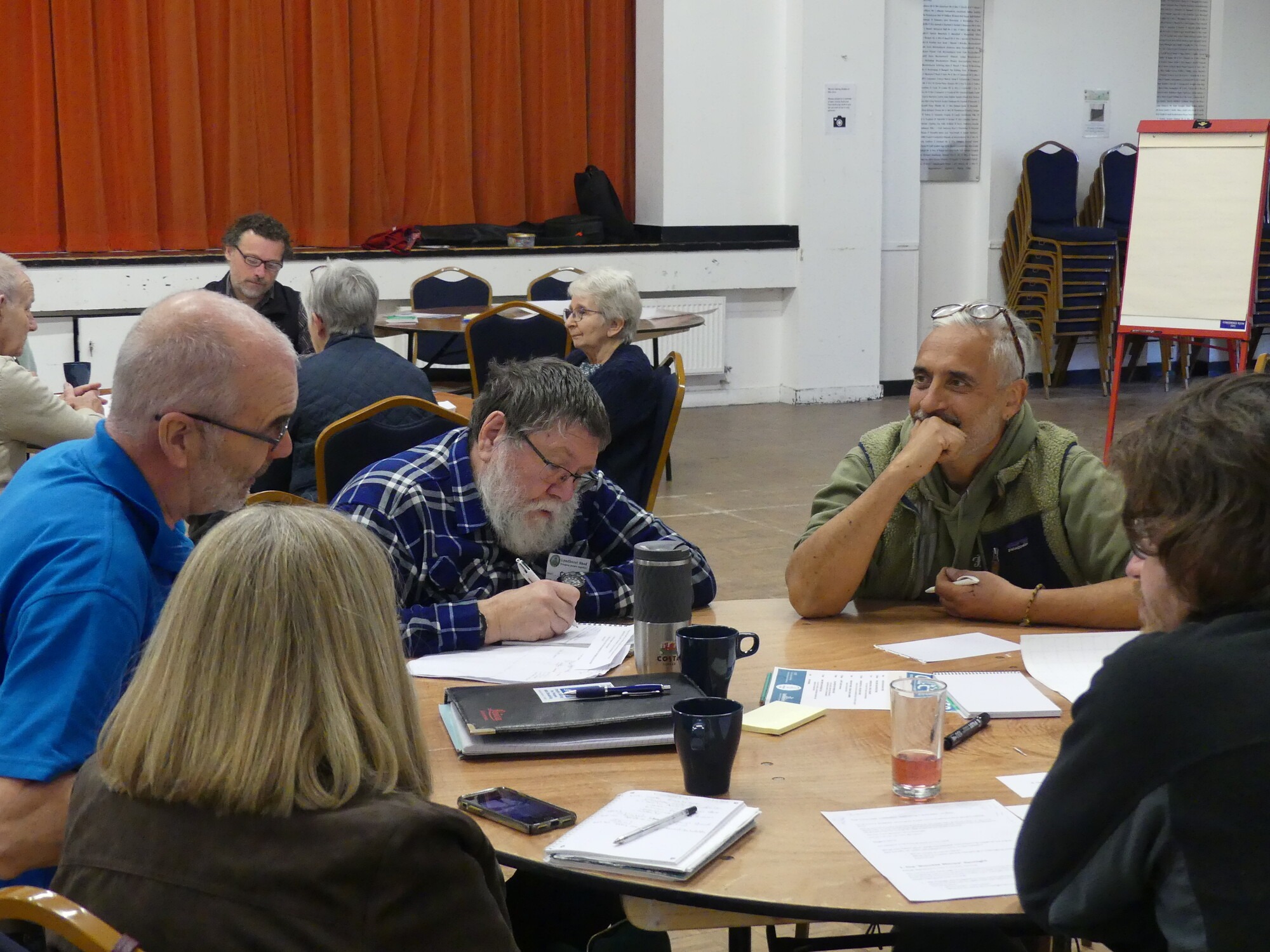 New Forest Volunteer Collective meeting, people seated at tables discussing and taking notes in a hall with orange stage curtain