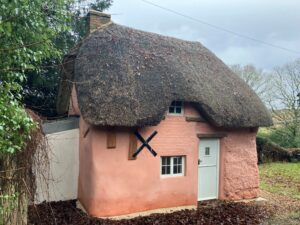 Pink thatched-roof cottage with white door and small windows, brick chimney, and wooded countryside surroundings