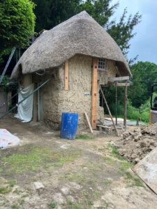 Thatched-roof cob cottage under restoration with scaffolding, timber supports, and construction materials in a yard