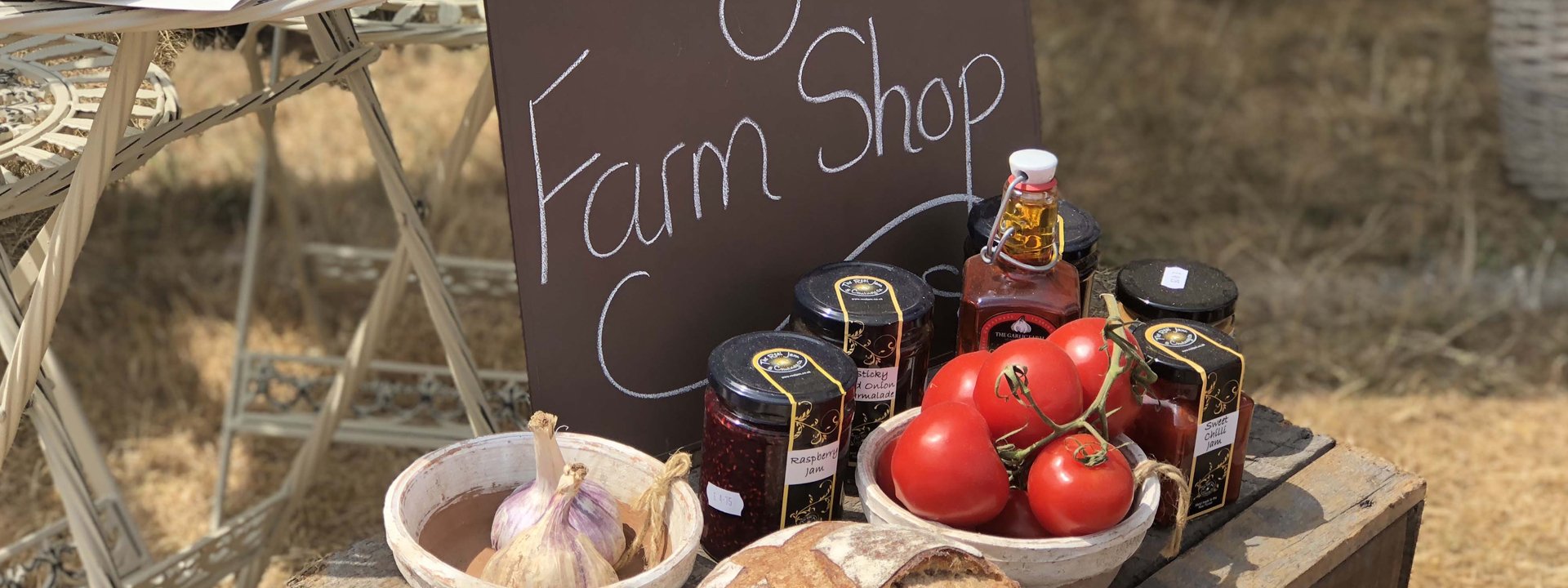 Farm Shop produce including tomatoes, jarred preserves and bowls of veg sit on a crate with a black board behind.