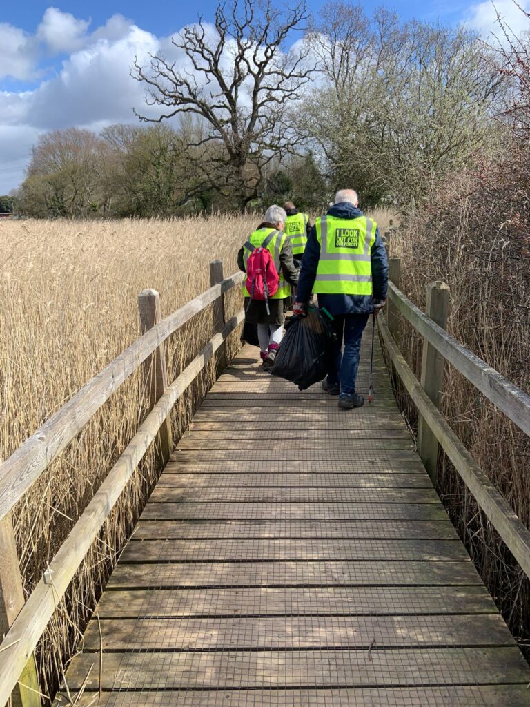 Three volunteers in hi-vis vests walk along a wooden boardwalk through reeds, carrying litter bags and pickers
