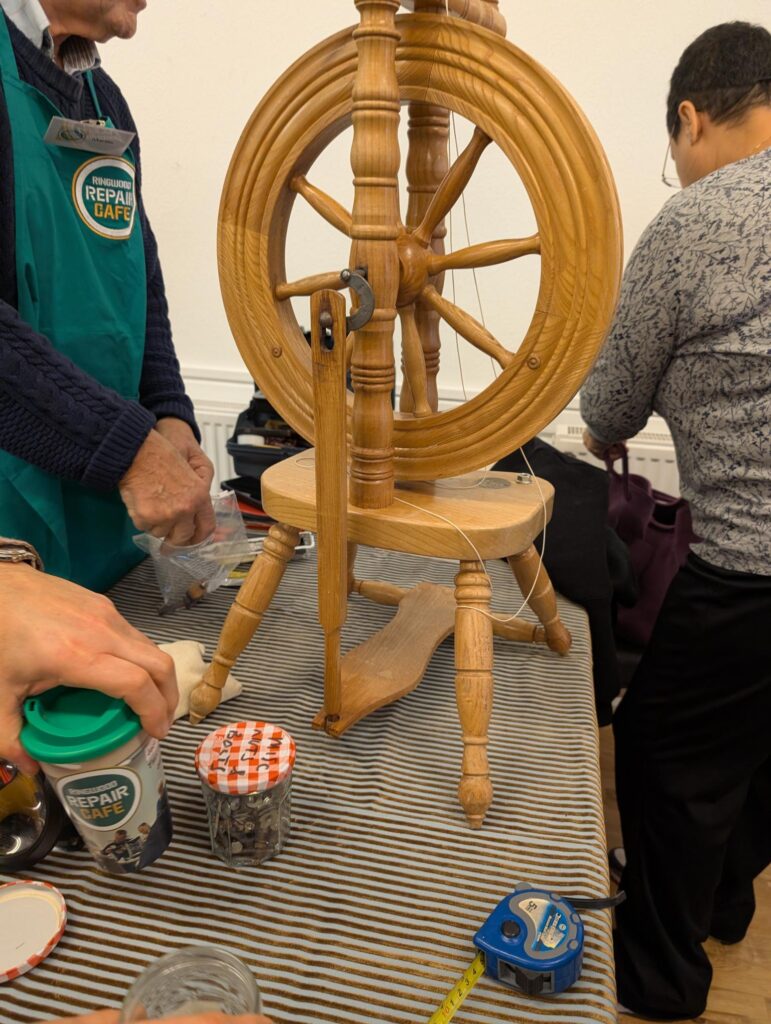 Old fashioned wooden spinning wheel in the centre of a table, being repaired and inspected by 2 people standing either side